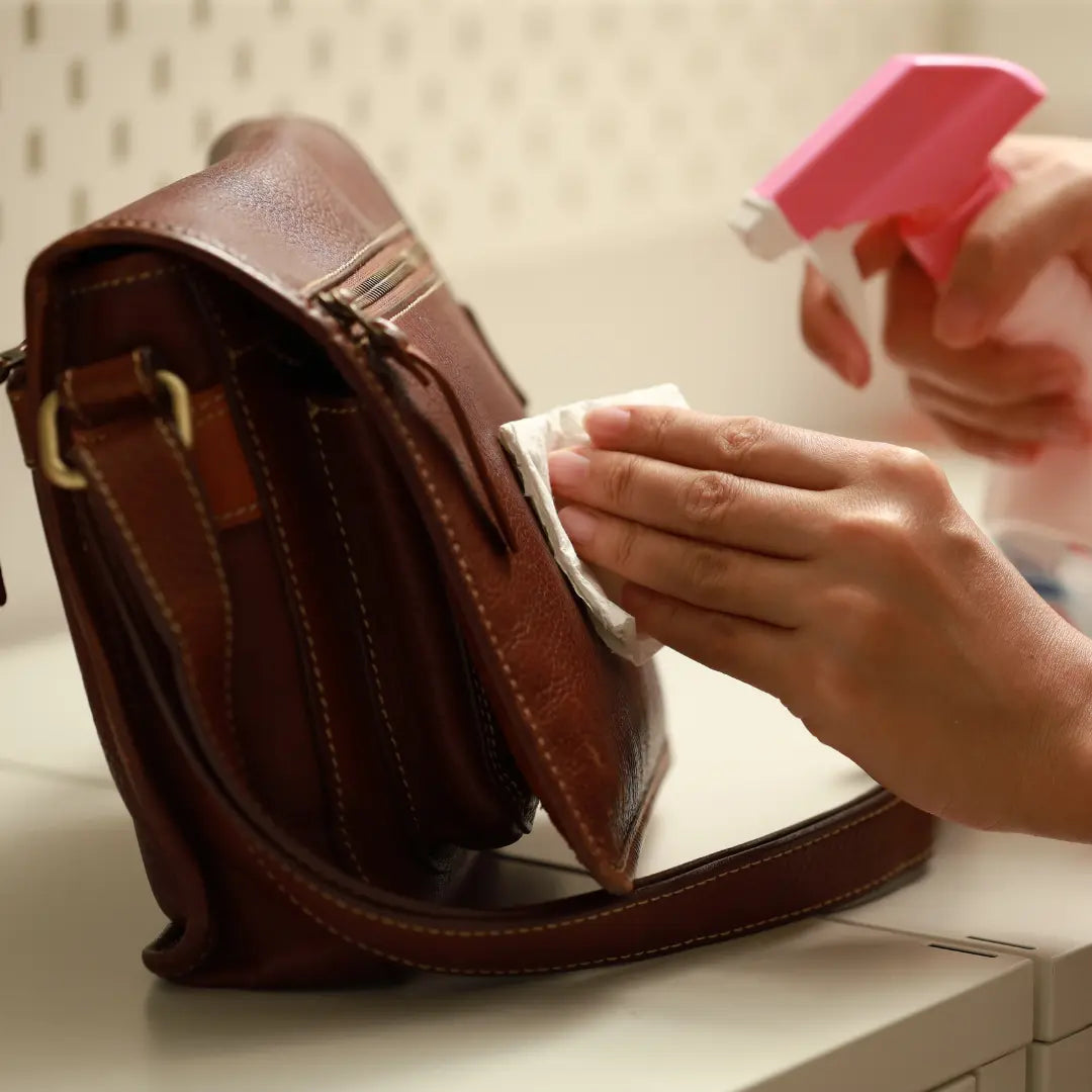 Persoon reinigt een bruine leren laptoptas met doekje en spray, toont hoe onderhoud je een leren laptoptas correct om de levensduur te verlengen.