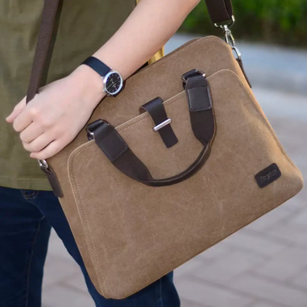Canvas laptoptas heren gedragen door persoon met horloge, lichtbruine tas met voorvak en praktische draaggreep.