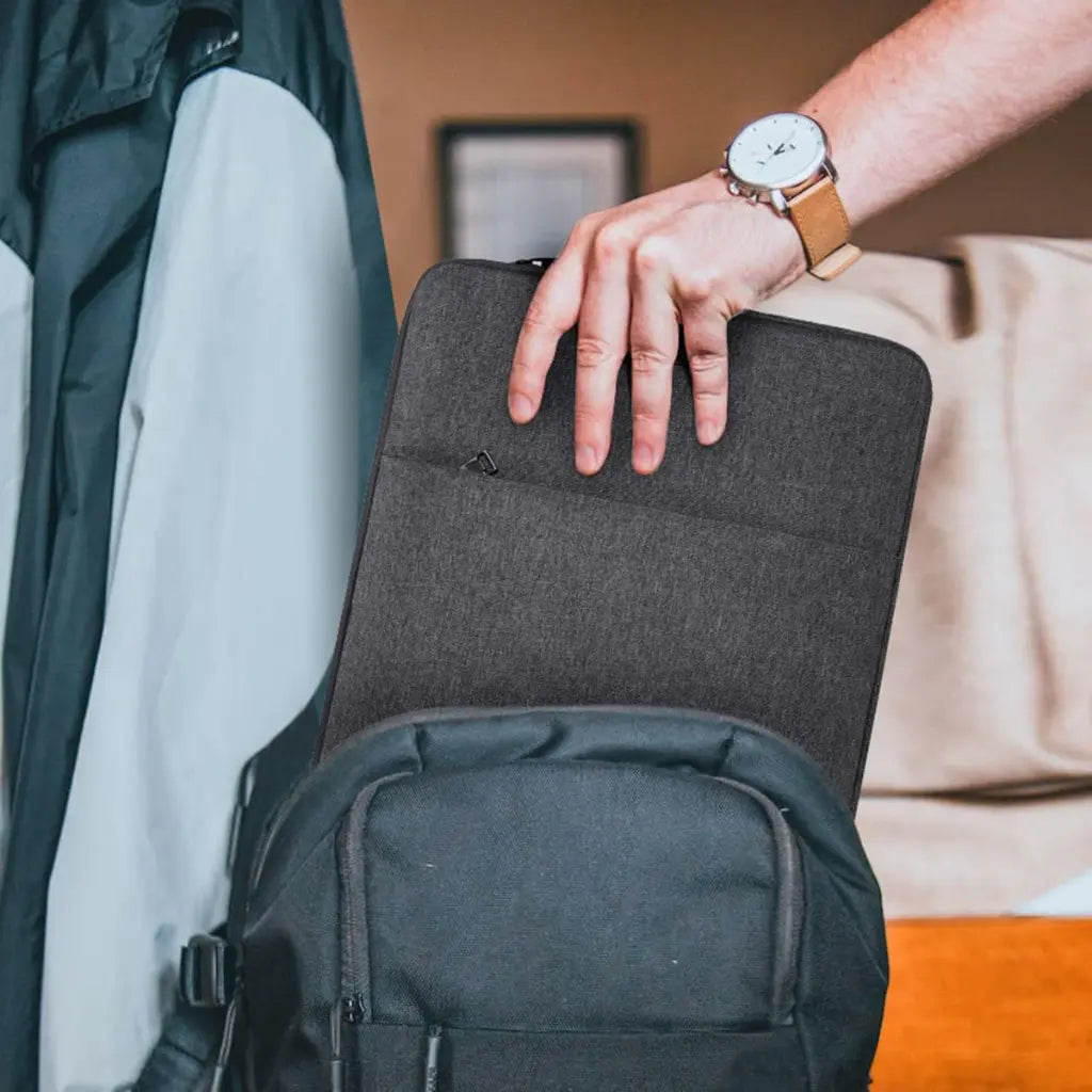 Hand met horloge die een laptop sleeve grey vasthoudt boven een rugzak.