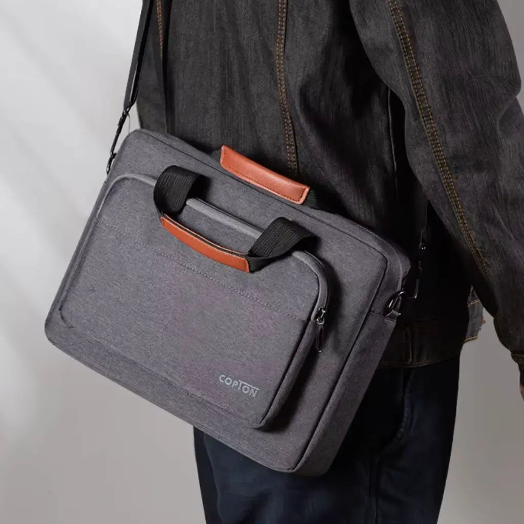 Design laptoptas heren donkergrijs gedragen over de schouder door een persoon in denimjas. De tas toont de bruine lederen handgreep en het praktische formaat, perfect voor dagelijks woon-werkverkeer en zakelijke bijeenkomsten.