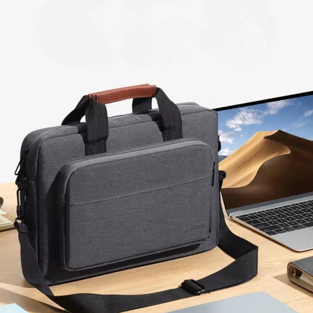 Design laptoptas heren donkergrijs naast een moderne laptop op een houten bureau. De elegante tas met bruin lederen handgreep en zwarte schouderband vormt een perfecte combinatie voor de moderne professional. Het COPTOT logo is subtiel zichtbaar op de voorkant.