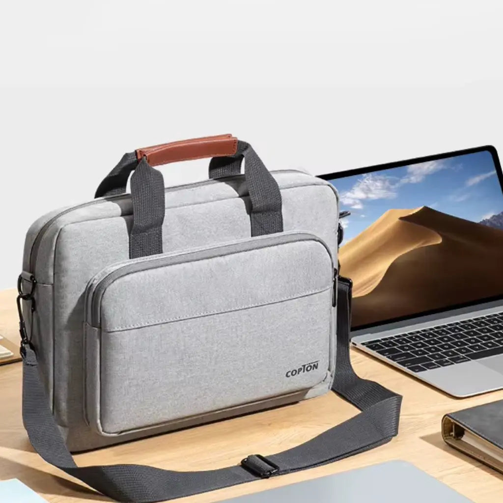 Design laptoptas heren lichtgrijs op een houten bureau naast een laptop. De COPTON tas met bruin lederen handgreep en zwarte schouderband is perfect gepositioneerd in een professionele werkomgeving, klaar voor dagelijks gebruik.