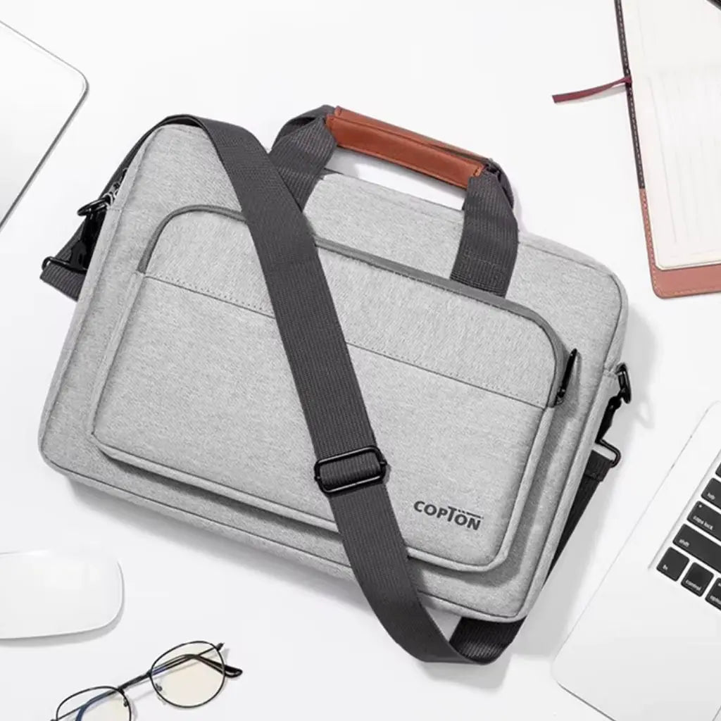 Design laptoptas heren lichtgrijs op een bureau naast een laptop en kantoorartikelen. De functionele tas met bruin lederen handgreep en zwarte schouderband biedt ruimte voor laptops en accessoires in een stijlvolle lichtgrijze uitvoering met het COPTON logo.