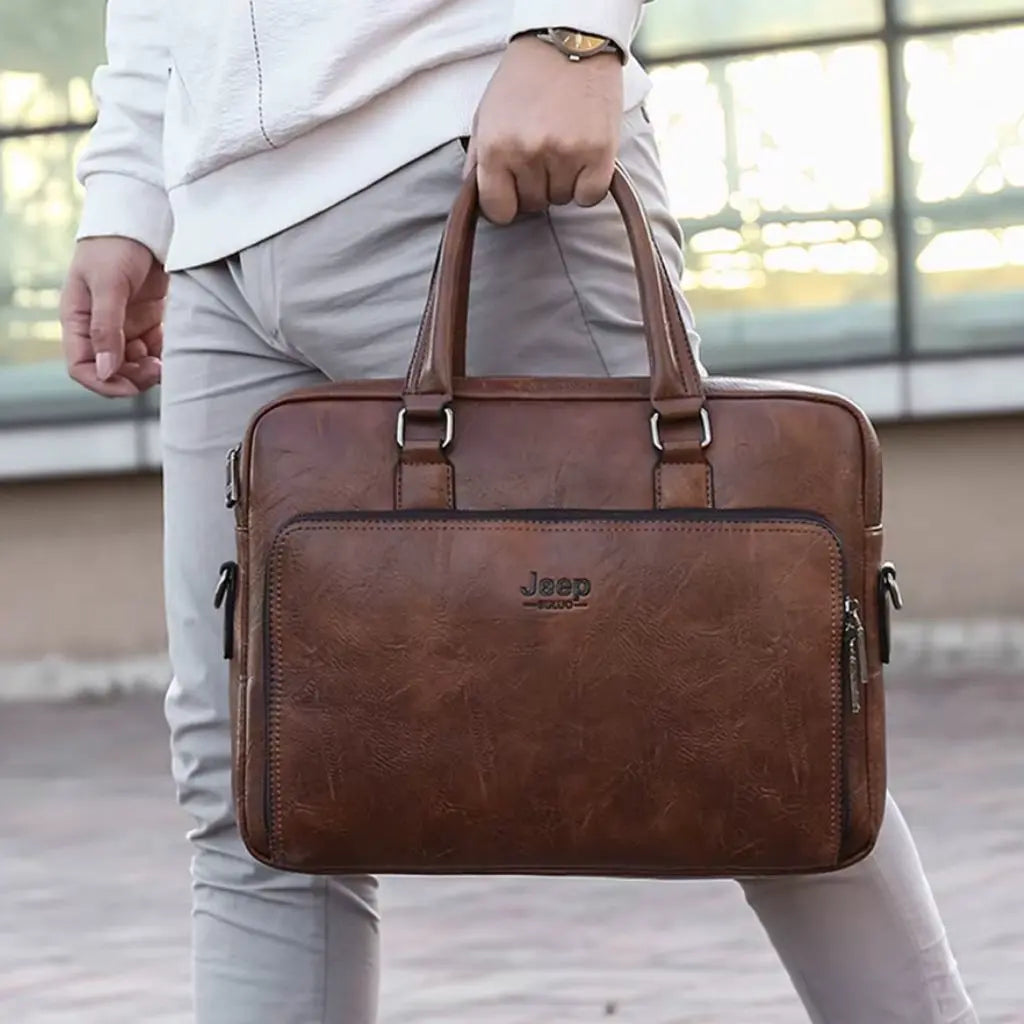 Exclusieve laptoptas heren 14 inch bruin vastgehouden aan de handgreep door een man met wit overhemd en horloge. De cognackleurige laptoptas toont het Jeep-logo, de functionele voorzakken en de hoogwaardige afwerking van het kunstleer voor dagelijks professioneel gebruik.