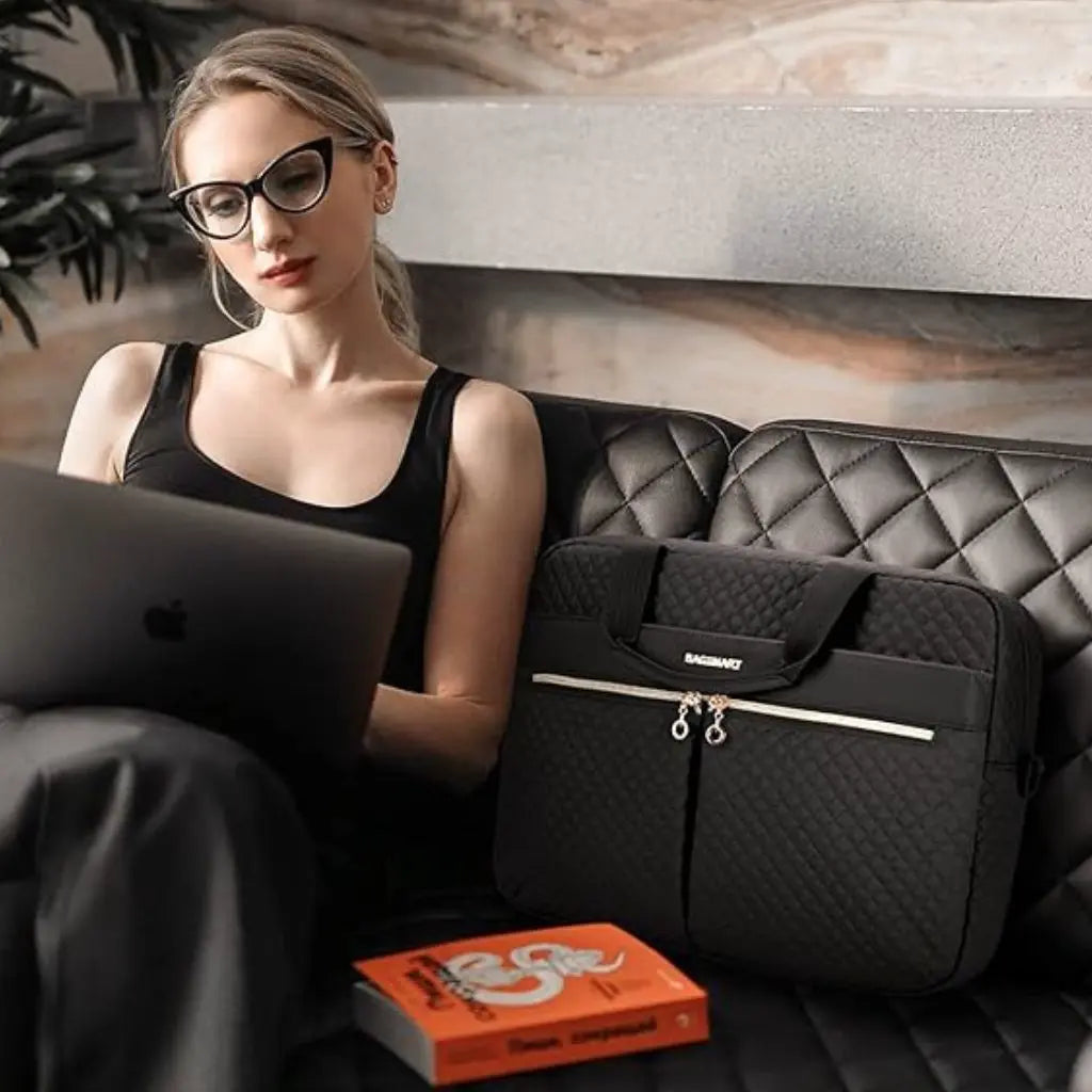Jonge vrouw met bril gebruikt laptop naast haar hippe laptoptas dames 17 inch zwart. De elegante BAGSMART tas rust op een luxe leren bank, toont het gestoffeerde ontwerp en gouden accenten perfect voor professioneel gebruik.