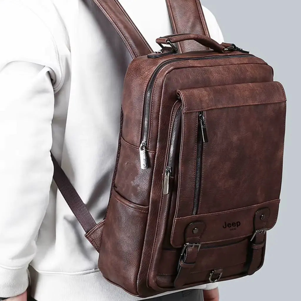 Man in witte trui met een leren laptop rugzak 15.6 inch bruin, met meerdere ritsvakken en een stevige leren handgreep.