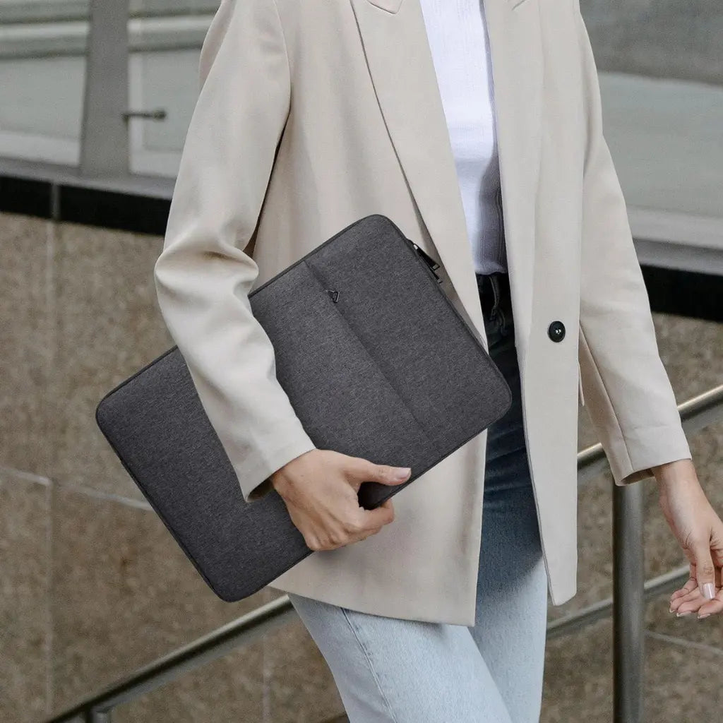 Vrouw in lichte jas die een laptop sleeve grey vasthoudt terwijl ze loopt.