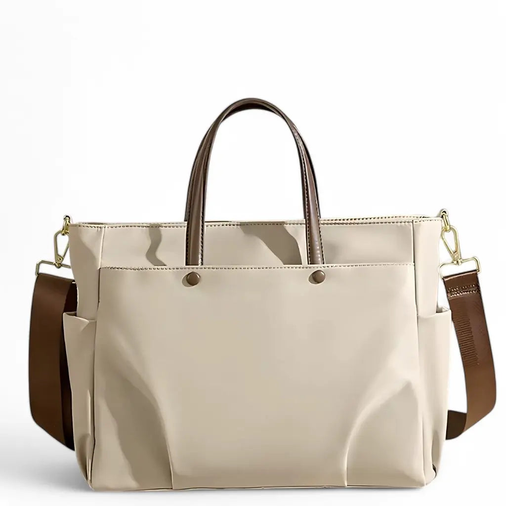 Een elegante beige Trendy laptoptas dames 15.6 inch met bruine lederen handgrepen en een verstelbare schouderriem. De moderne laptoptas dames 15.6 inch heeft een gestroomlijnd ontwerp met metalen knopen en zijvakken voor extra opbergruimte, ideaal voor professioneel gebruik.