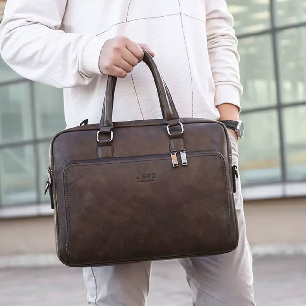 Close-up van de exclusieve laptoptas heren 14 inch donkerbruin vastgehouden aan de handgreep, met zichtbaar Jeep-logo en dubbele ritssluiting voor extra veiligheid.