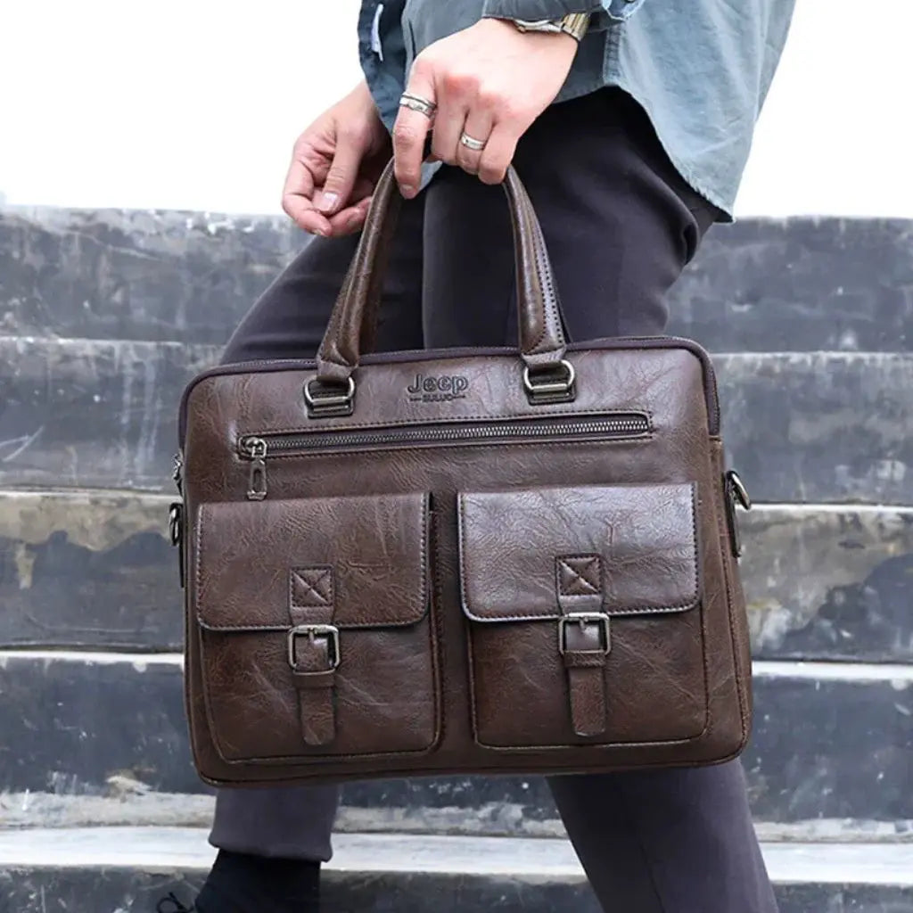 Close-up van een persoon die de leren laptoptas heren 13 inch bruin donkerbruin vasthoudt aan de handgreep, met duidelijk zichtbaar Jeep logo en robuuste vakken met metalen gespen.
