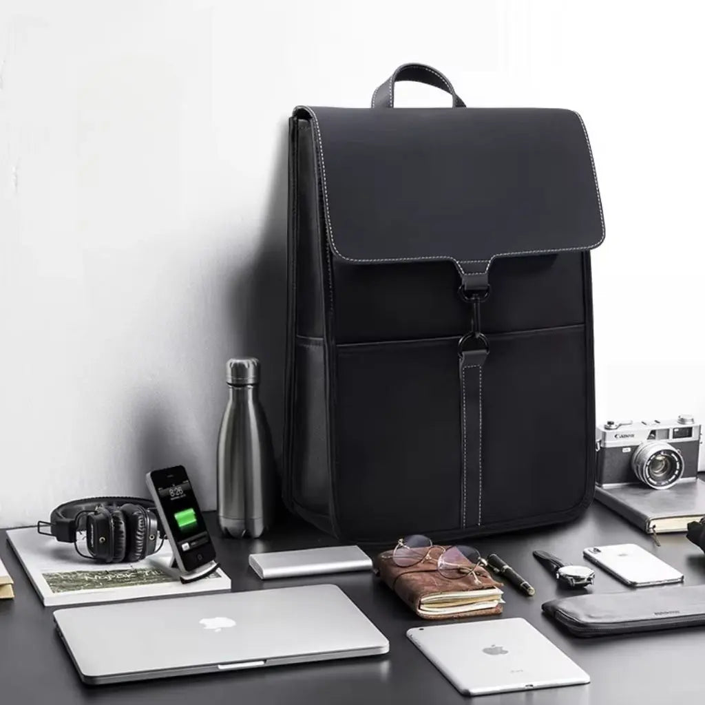 Elegante zwarte rugzak voor laptop van 15,6 inch, geplaatst op een bureau met verschillende accessoires zoals een MacBook, iPad, smartphone, notitieboek, bril en een waterfles.