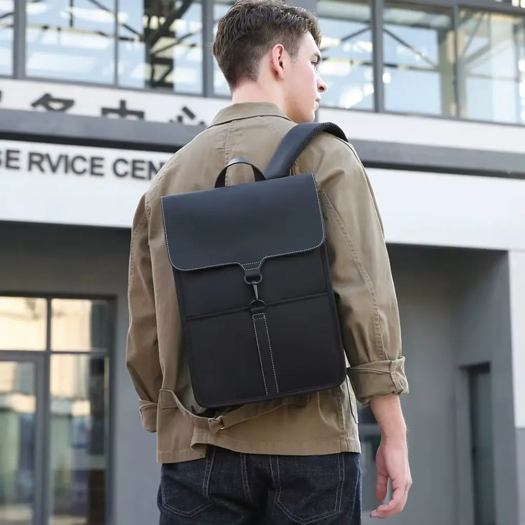 Jonge man met een stijlvolle zwarte rugzak voor laptop 15,6 inch, gedragen op zijn rug. Hij staat buiten voor een modern gebouw en straalt een modieuze en zakelijke uitstraling uit.