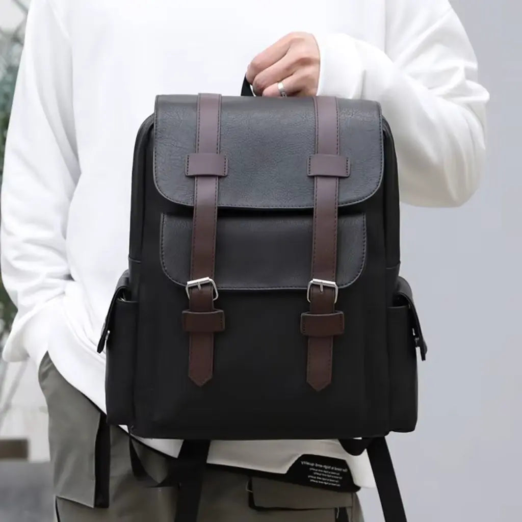 Een persoon draagt de Heren laptop rugzak 13 inch zwart in de hand, met een stijlvol ontwerp en bruine lederen gespen voor een vintage uitstraling.