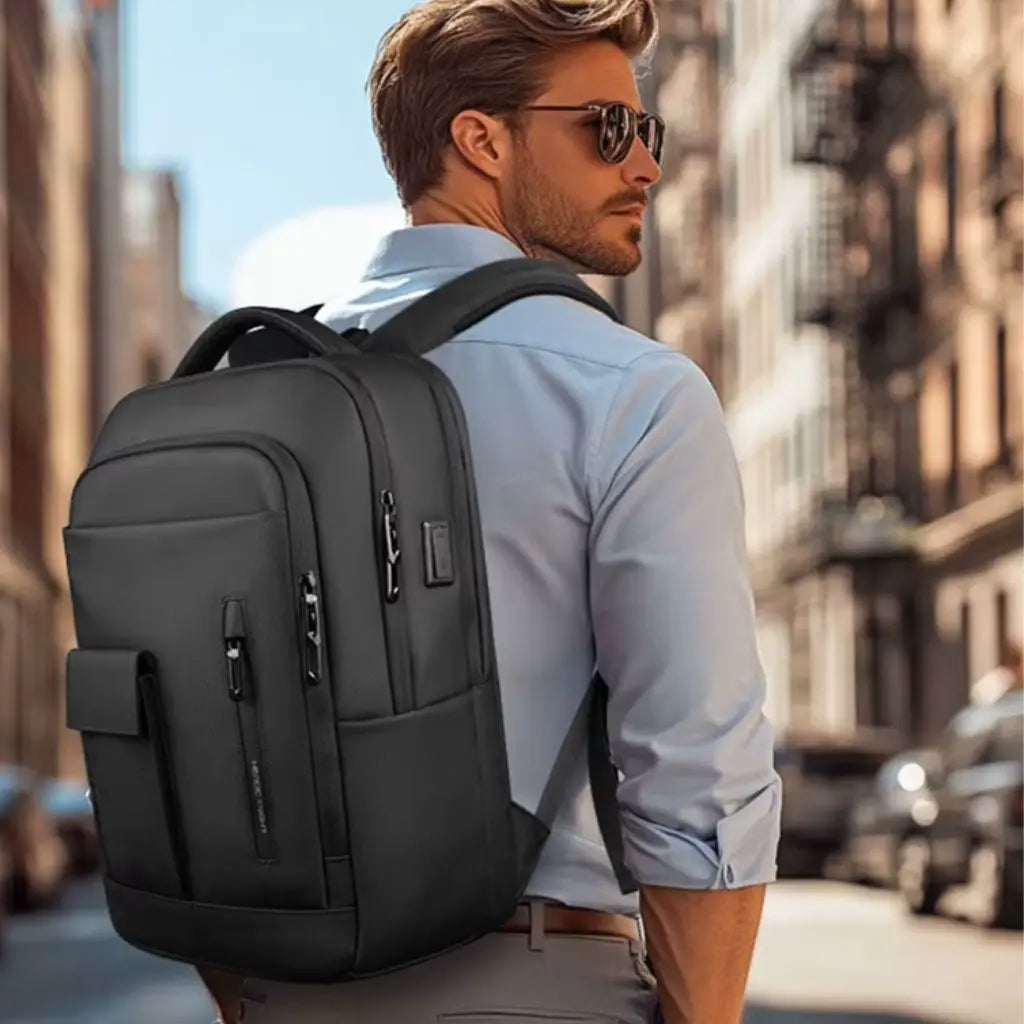 Elegante man met zonnebril en een laptop rugzak 16 inch zwart op de rug, wandelend door een drukke stad.