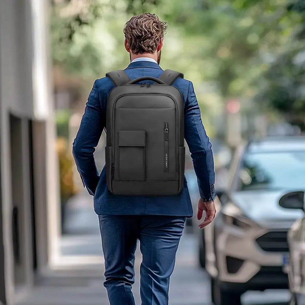 Man in een stijlvol blauw pak draagt een laptop rugzak 16 inch zwart terwijl hij door een stedelijke omgeving loopt.