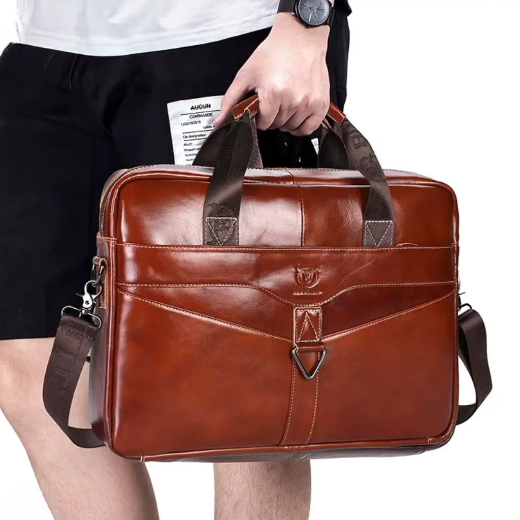 Persoon in zwarte broek houdt laptoptas cognac heren 15 inch vast aan de handgreep met documenten in de andere hand.
