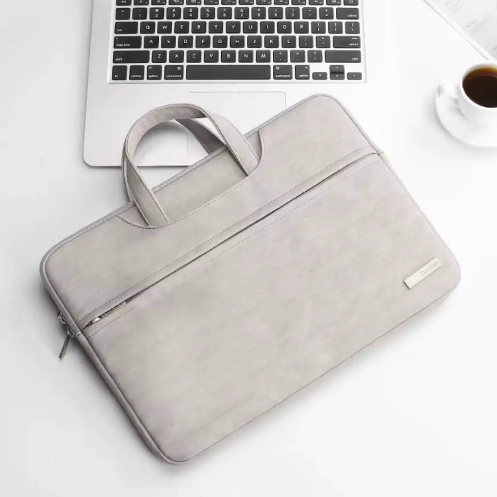 Laptoptas dames klein grijs naast een MacBook laptop en koffiekopje op een witte bureau-achtergrond. De functionele tas toont de praktische handgrepen en het voorvak met ritssluiting, ideaal voor professionele vrouwen die onderweg werken.