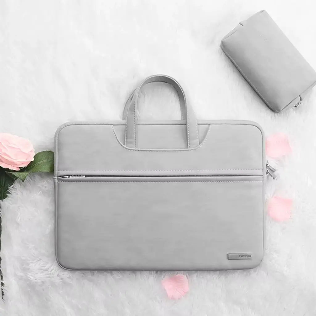 Laptoptas dames klein grijs gepresenteerd op een zachte witte ondergrond met roze bloemblaadjes en een bijpassende accessoire-tas. Het elegante grijze design met TAIKESEN-label vormt een stijlvolle combinatie voor vrouwen die functionaliteit en mode waarderen.