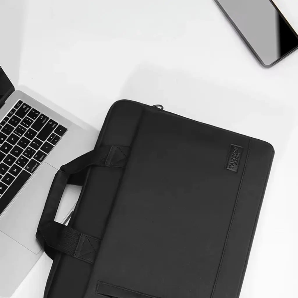 Bovenaanzicht van een minimalistisch wit bureau met de laptoptas heren 17 inch zwart naast een MacBook laptop en smartphone. De compacte TAIKESEN tas met handgrepen is een perfect accessoire voor de moderne professional in een strakke werkomgeving.