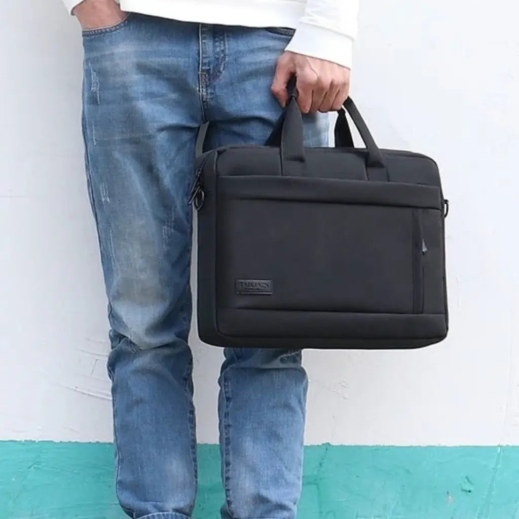 Man in casual kleding met witte trui en spijkerbroek houdt de laptoptas heren 17 inch zwart vast tegen een witte muur. De elegante zwarte TAIKESEN tas toont het ruime ontwerp met handgrepen, perfect voor dagelijks gebruik in een informele setting.