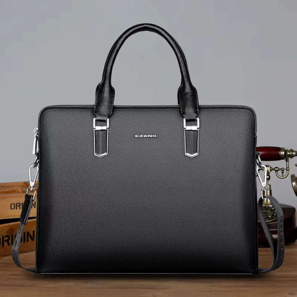 Een nette laptoptas heren 14 inch zwart op een houten bureau met het KOMANSO logo, vervaardigd uit premium kunstleder met karakteristieke textuur, ergonomische handgrepen en duurzame metalen bevestigingspunten voor de schouderriem.