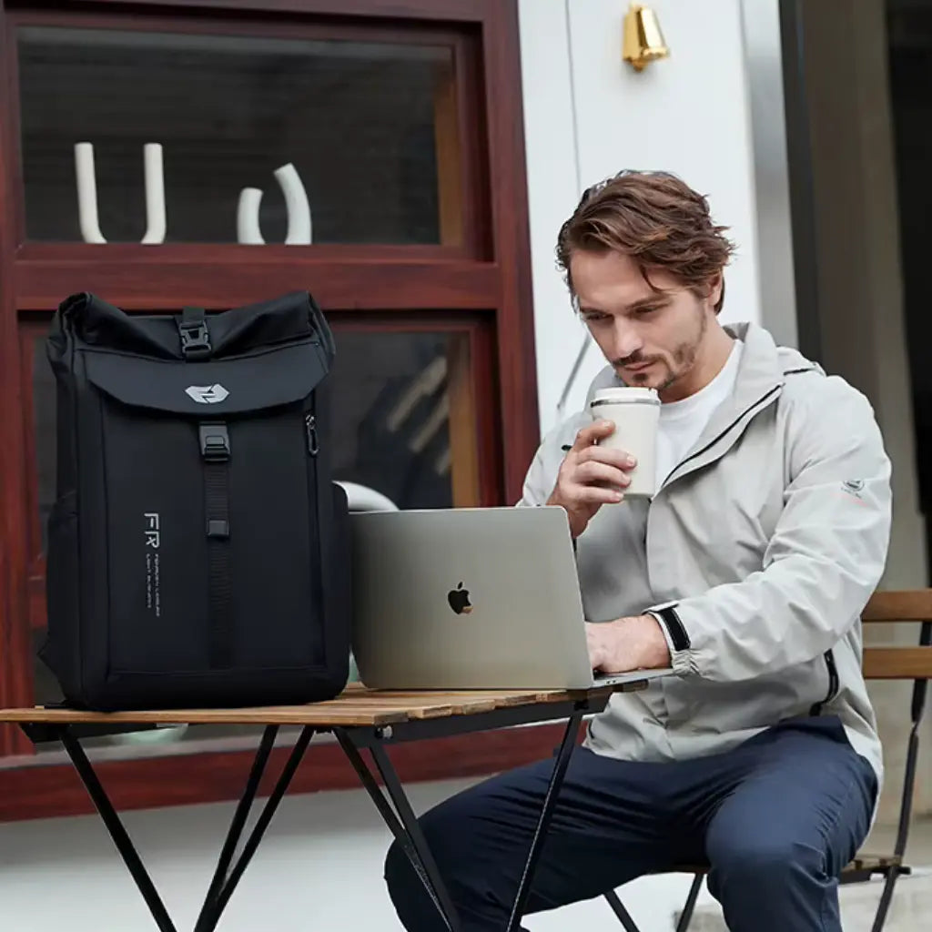 Man werkt op MacBook op buitenterras met koffie naast zijn zwarte Nomad laptop rugzak, perfect voor digitale nomaden.