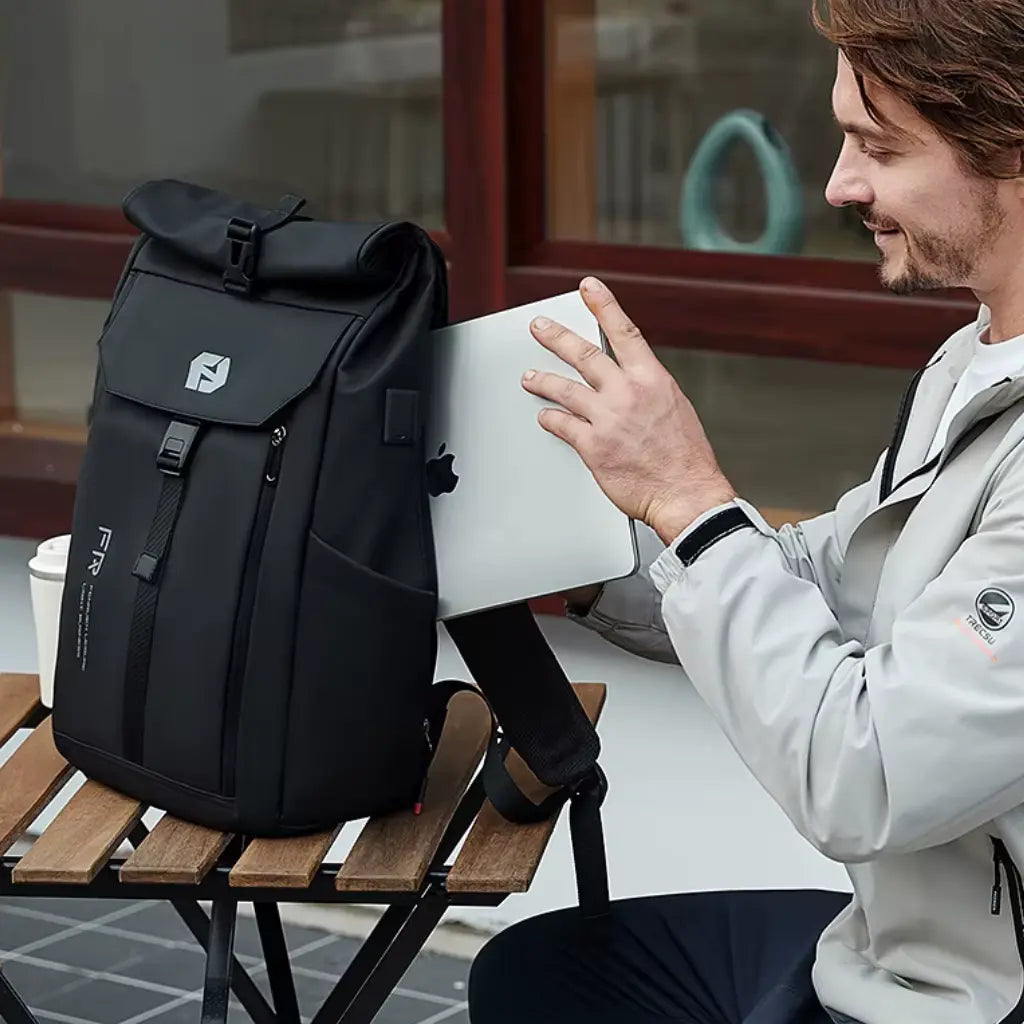 Man in lichte jas zit op terras met MacBook naast zwarte Nomad laptop rugzak, ideaal voor professionals onderweg.