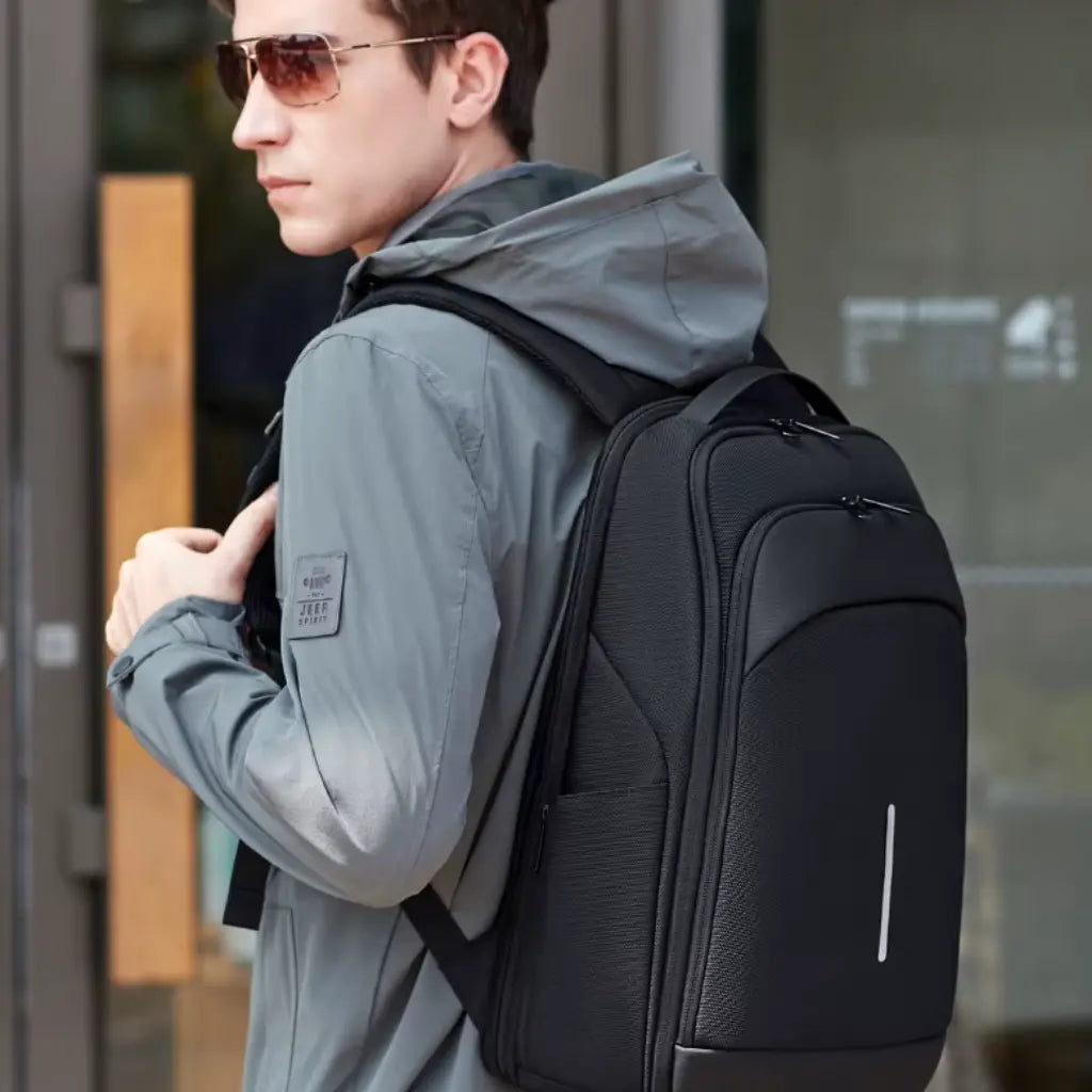 Jonge man met zonnebril draagt waterdichte laptop rugzak school 16 inch met lichtgrijze jas, perfect voor dagelijks kantoorgebruik en casual zakelijke setting.