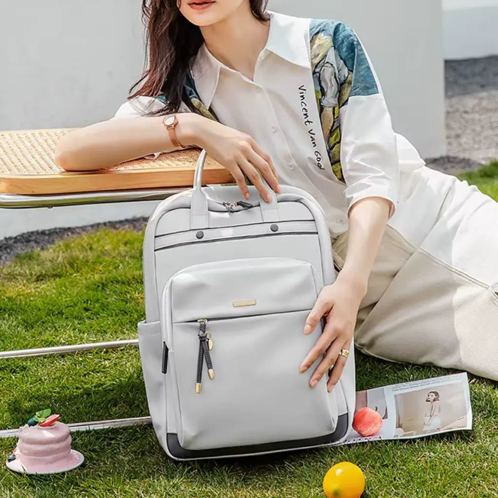Een elegante waterdichte laptop rugzak 15.6 inch grijs geplaatst op een grasveld tijdens een buitenpicknick. Een jonge vrouw in een witte blouse met artistieke details zit naast de rugzak. De grijze tas heeft een stijlvol ontwerp met voorvak, metalen knoppen en decoratieve hangers, perfect voor een dag buiten terwijl je laptop veilig blijft.