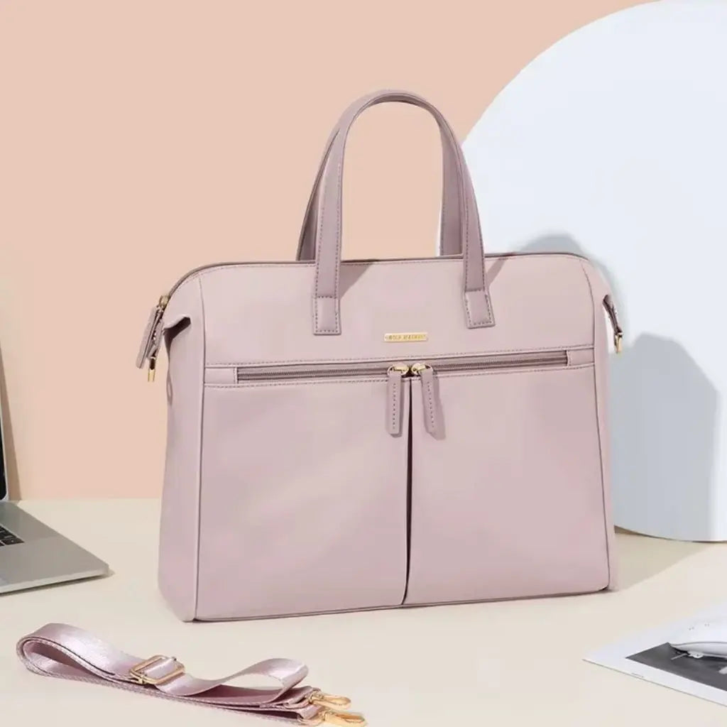 Een stijlvolle roze laptoptas dames 15.6 inch met afneembare schouderriem op een bureau naast een laptopcomputer. De laptoptas dames 15.6 inch roze past perfect in een moderne werksfeer en kan zowel als hand- als schoudertas worden gedragen.